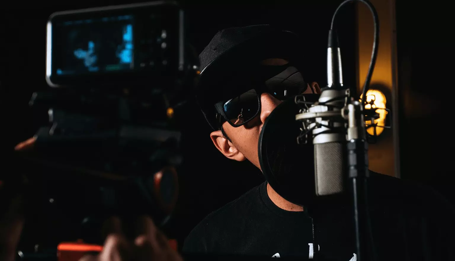man with cap and sunglasses rapping in hanging microphone with a decent yellow light in the background